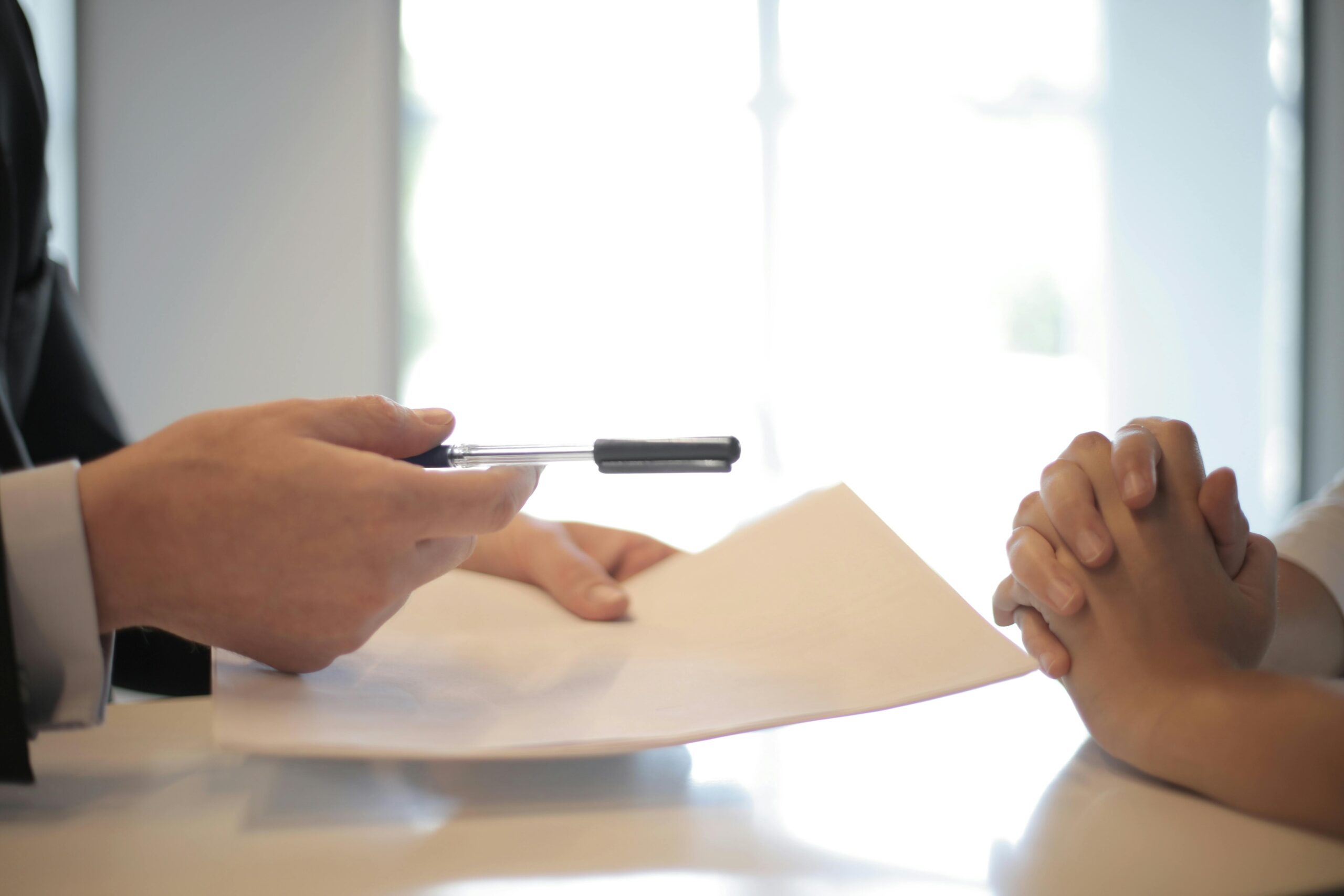 Une personne tend un stylo et un document à une autre personne, illustrant une situation de signature ou de discussion contractuelle dans un cadre professionnel